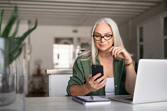 Mulher de óculos olhando o celular de frente para o computador