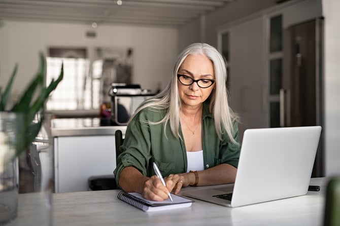 Mulher com cabelo branco usando óculos e uma camisa verde militar escrevendo no caderno enquanto está com o laptop aberto para ver se a Conta Mercado Pago é só para quem vende