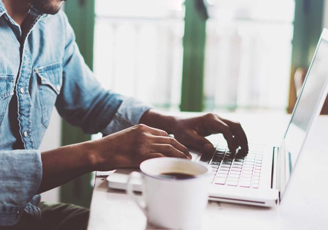 Homem mexendo no laptop em uma mesa com uma xícara de café ao lado