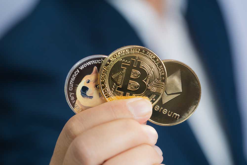 Uma mão segurando três moedas douradas com os símbolos da Dogecoin, Bitcoin e Ethereum. 