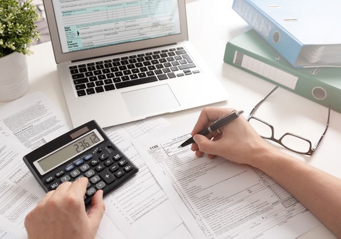 Mercado Pago: mãos de empreendedor em mesa com notebook,calculadora e papéis organizando os documentos para declarar o imposto de renda. Na mesa há pastas e um vaso de planta.