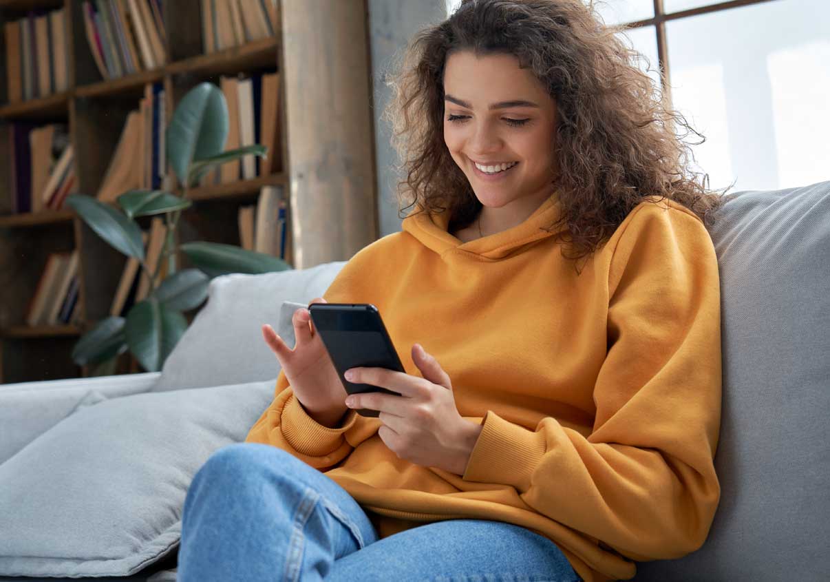Uma adolescente sentada em um sofá, sorrindo e mexendo em um celular