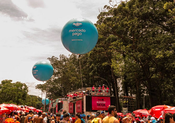 Mercado Pago: Imagem de foliões comemorando o Carnaval de Rua em uma avenida cercada por árvores. No centro há dois trios elétricos e dois balões com o logo do Mercado Pago.